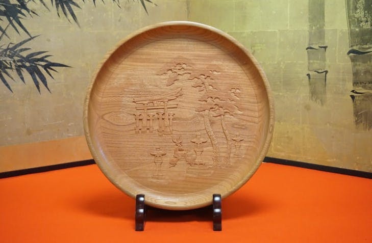 A round wooden tray with an engraved design featuring a traditional Japanese torii gate, trees, and lanterns, displayed upright on a black stand against an orange and beige background with bamboo patterns.
