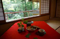 A traditional Japanese tea set with sweets sits on a red cloth inside a tatami room, with sliding shoji doors open to a lush green garden outside. Maple leaves decorate the table.