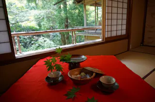A traditional Japanese tea set with sweets sits on a red cloth inside a tatami room, with sliding shoji doors open to a lush green garden outside. Maple leaves decorate the table.