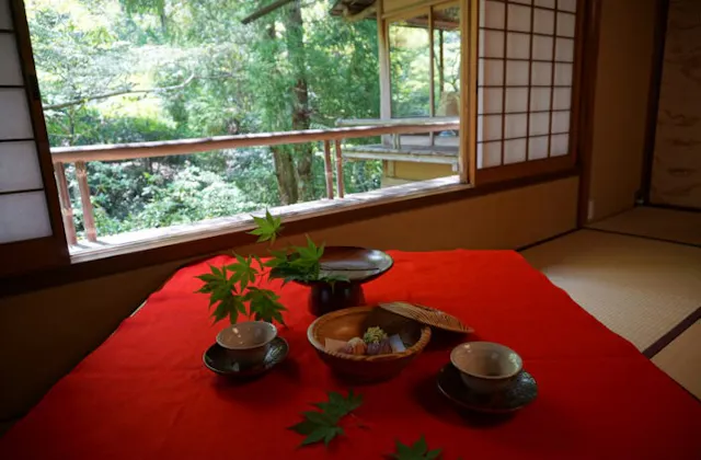A traditional Japanese tea set with sweets sits on a red cloth inside a tatami room, with sliding shoji doors open to a lush green garden outside. Maple leaves decorate the table.