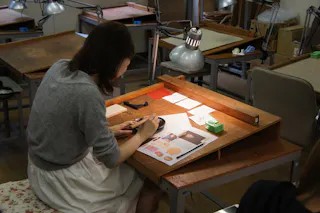 A woman sits at a wooden desk under a lamp, working with her hands on a small craft project. Papers, tools, and materials are spread out on the desk in a workshop setting with other empty desks nearby.