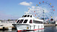 A white passenger boat is docked near a pier, with a colorful Ferris wheel and blue sky in the background. The water is calm and the scene appears to be at an amusement area or waterfront park.