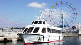 A white passenger boat is docked near a pier, with a colorful Ferris wheel and blue sky in the background. The water is calm and the scene appears to be at an amusement area or waterfront park.