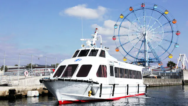 A white passenger boat is docked near a pier, with a colorful Ferris wheel and blue sky in the background. The water is calm and the scene appears to be at an amusement area or waterfront park.