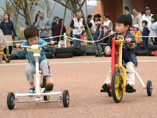 Two young boys ride tricycles on a playground while a crowd of adults and children watch in the background. The boys appear focused on their ride and the area is surrounded by rope and tires.