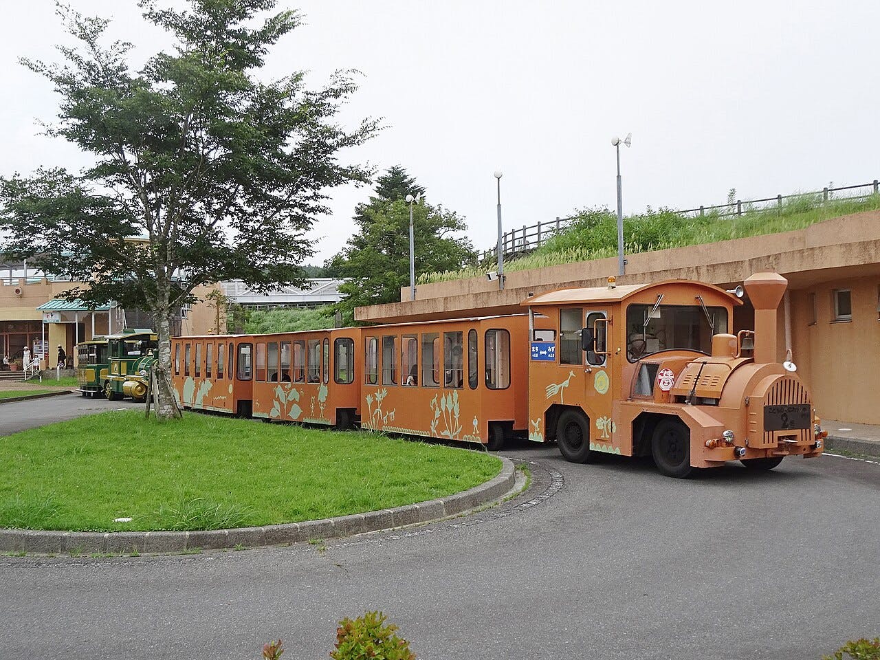 A small, orange, open-air tour train with animal designs on its sides travels along a circular paved path in a park-like area with grass, trees, and nearby buildings.