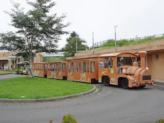 A small, orange, open-air tour train with animal designs on its sides travels along a circular paved path in a park-like area with grass, trees, and nearby buildings.
