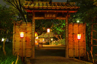 A traditional Japanese wooden gate with lanterns illuminating a garden path at night, bordered by bamboo fences and lush greenery.