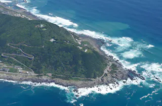 Aerial view of a green, rocky peninsula surrounded by blue ocean waves, with winding roads and scattered buildings visible along the coastline.