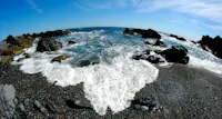 Waves crash against dark rocks and a pebbly beach under a clear blue sky, viewed with a fisheye lens that curves the horizon and distorts the scene, creating a dynamic, wide-angle perspective of the coastline.