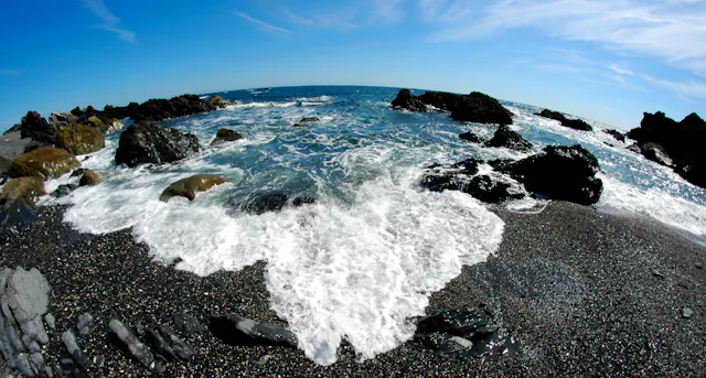Waves crash against dark rocks and a pebbly beach under a clear blue sky, viewed with a fisheye lens that curves the horizon and distorts the scene, creating a dynamic, wide-angle perspective of the coastline.