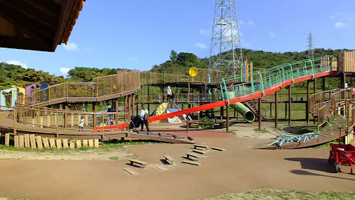 Large playground with various climbing structures, slides, and bridges on a sunny day. Children are playing, and there is green grass, trees, and hills in the background. A tall utility tower is visible behind the playground.