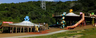 An outdoor playground with large spiral and tube slides, elevated platforms, and whimsical structures, surrounded by lush green trees and hills under a clear blue sky.