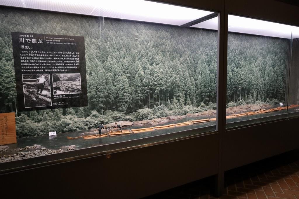 A glass display case features a diorama of a forest with a river, wooden logs floating in the water, and small human figurines depicting log transportation; Japanese text and photos are mounted on the back panel.