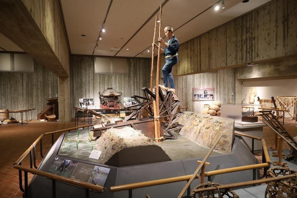 A museum exhibit featuring a life-sized mannequin of a person standing on a wooden structure, with surrounding displays of traditional tools, equipment, and informational signs in a spacious, well-lit room.