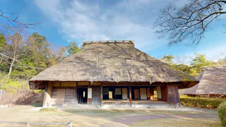 A traditional Japanese farmhouse with a large thatched roof, wooden walls, and sliding doors, surrounded by trees and under a blue sky.
