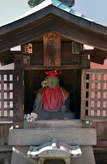 A stone statue dressed in a red bib and hat sits inside a small wooden shrine with a sloped roof, lattice windows, and Japanese inscriptions. A white artificial flower rests at the statue’s base.