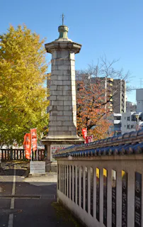 A tall stone monument stands beside a tiled wall engraved with Japanese characters, surrounded by autumn trees and red banners, with modern city buildings in the background under a clear blue sky.