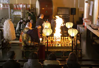 A Buddhist monk performs a fire ritual before an altar with flames rising, surrounded by lanterns and offerings, as several people watch from the audience in a dimly lit indoor setting.