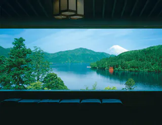 A large window frames a scenic view of a serene lake, lush green trees, distant mountains, and Mount Fuji with a snow-capped peak under a clear blue sky. A red torii gate stands by the shore.