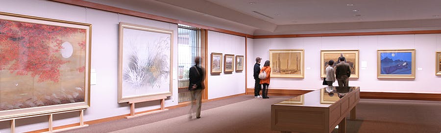 People admire paintings in a spacious, well-lit art gallery. Large framed artworks hang on the pale walls and a glass display case is at the center. The atmosphere is calm and reflective.