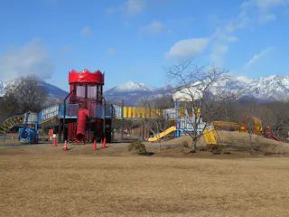 A colorful playground with slides, towers, and climbing structures stands on a dry grassy field. Snow-capped mountains and leafless trees are visible in the background under a blue sky.