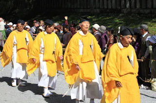 Four men wearing traditional yellow and white Japanese garments and black caps walk in a procession, with a crowd of onlookers standing behind them outdoors on a sunny day.