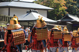 A group of people wearing traditional Japanese samurai armor with gold helmets and red details stand in a line outdoors, with onlookers and traditional buildings in the background.