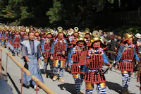 A group of people in traditional samurai armor, with gold helmets and red, black, and blue attire, march in a parade outdoors, watched by a crowd behind wooden barriers. Trees and sunlight are in the background.