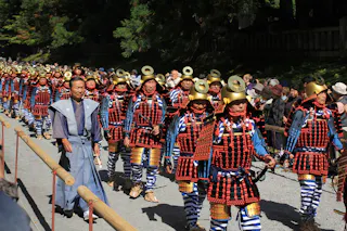 A group of people in traditional samurai armor, with gold helmets and red, black, and blue attire, march in a parade outdoors, watched by a crowd behind wooden barriers. Trees and sunlight are in the background.