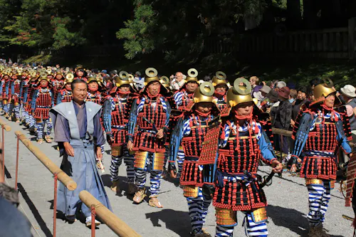 A group of people in traditional samurai armor, with gold helmets and red, black, and blue attire, march in a parade outdoors, watched by a crowd behind wooden barriers. Trees and sunlight are in the background.