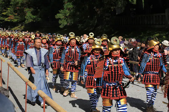 A group of people in traditional samurai armor, with gold helmets and red, black, and blue attire, march in a parade outdoors, watched by a crowd behind wooden barriers. Trees and sunlight are in the background.