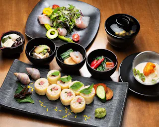 A beautifully arranged Japanese meal featuring assorted sushi rolls, fresh salad, sea snails, soup, pickled vegetables, and dessert, all served in elegant black dishes on a wooden table.
