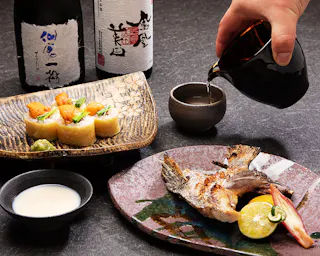 A hand pours sake into a small cup beside bottles of sake. On the table are a grilled fish dish with lime, tofu rolls topped with garnish, and another small cup of drink, all arranged on decorative plates.