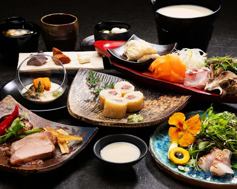 A variety of colorful Japanese dishes are artfully arranged on plates, featuring sushi rolls, sliced meat, fresh sashimi, vegetables, garnishes, soup, and a floral salad, all displayed on a dark tabletop.