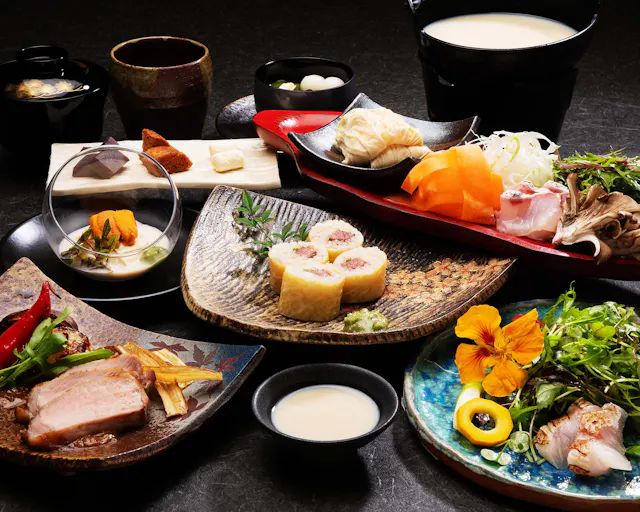 A variety of colorful Japanese dishes are artfully arranged on plates, featuring sushi rolls, sliced meat, fresh sashimi, vegetables, garnishes, soup, and a floral salad, all displayed on a dark tabletop.
