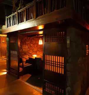 A cozy, dimly lit dining booth with wooden sliding doors and stone walls, illuminated by a warm hanging lantern, creates an inviting, rustic atmosphere.