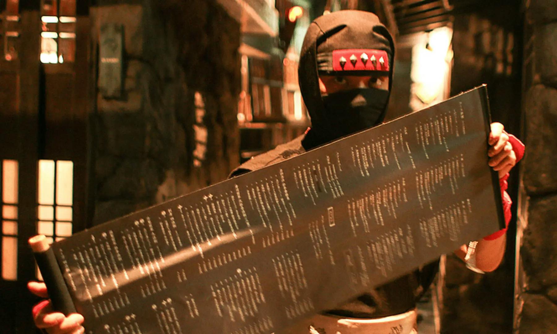 A person dressed in a black ninja costume holds a large, rectangular menu with rows of text in a dimly lit restaurant with stone walls.