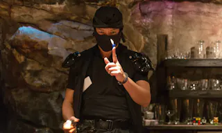 A person dressed in black ninja-style clothing holds a lighter with a small flame near their finger, standing in front of a rocky wall and shelves with glassware.