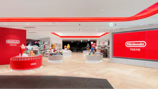 A brightly lit Nintendo Tokyo store with red walls, large Nintendo Tokyo signs, and displays featuring merchandise and character statues like Mario and Luigi. Shelves of products are arranged throughout the spacious interior.