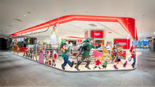 A brightly lit Nintendo store with large glass windows decorated with colorful images of popular Nintendo characters, such as Mario, Luigi, Link, and Kirby. Inside, shelves display various merchandise and Nintendo signage.