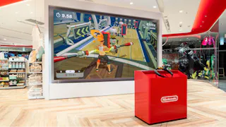 A large screen in a bright, modern store displays someone playing Nintendo's Ring Fit Adventure. A red Nintendo kiosk with game controllers is positioned in front of the screen. Shelves of merchandise are seen on the left.