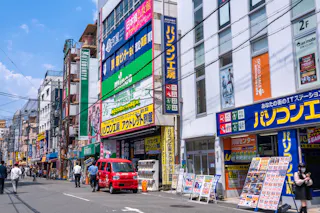 A busy street in Japan with colorful signs in Japanese, a red van parked by the sidewalk, pedestrians walking, and tall buildings lined with electronics and retail stores.