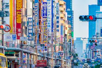 A busy city street in Japan filled with colorful vertical signs in Japanese and English, traffic lights, and power lines, with tall buildings in the background.