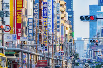 A busy city street in Japan filled with colorful vertical signs in Japanese and English, traffic lights, and power lines, with tall buildings in the background.