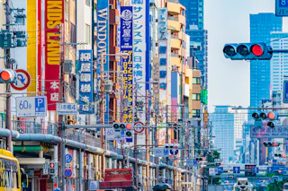 A busy city street in Japan filled with colorful vertical signs in Japanese and English, traffic lights, and power lines, with tall buildings in the background.