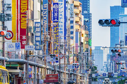 A busy city street in Japan filled with colorful vertical signs in Japanese and English, traffic lights, and power lines, with tall buildings in the background.