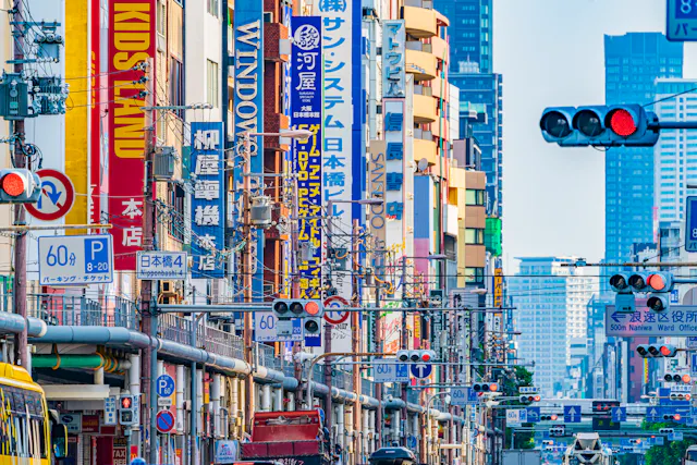 A busy city street in Japan filled with colorful vertical signs in Japanese and English, traffic lights, and power lines, with tall buildings in the background.