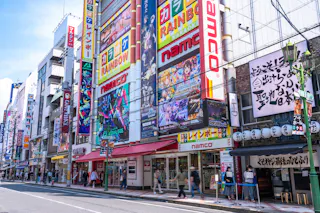 Colorful storefronts in Akihabara, Tokyo, featuring bright signage for anime, manga, and game shops, including Namco. People stroll along the street, and the buildings are covered with large posters and advertisements.