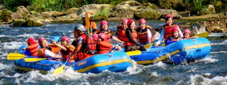 A group of people wearing helmets and life jackets enjoy white-water rafting in two blue inflatable rafts on a river surrounded by rocks and greenery.